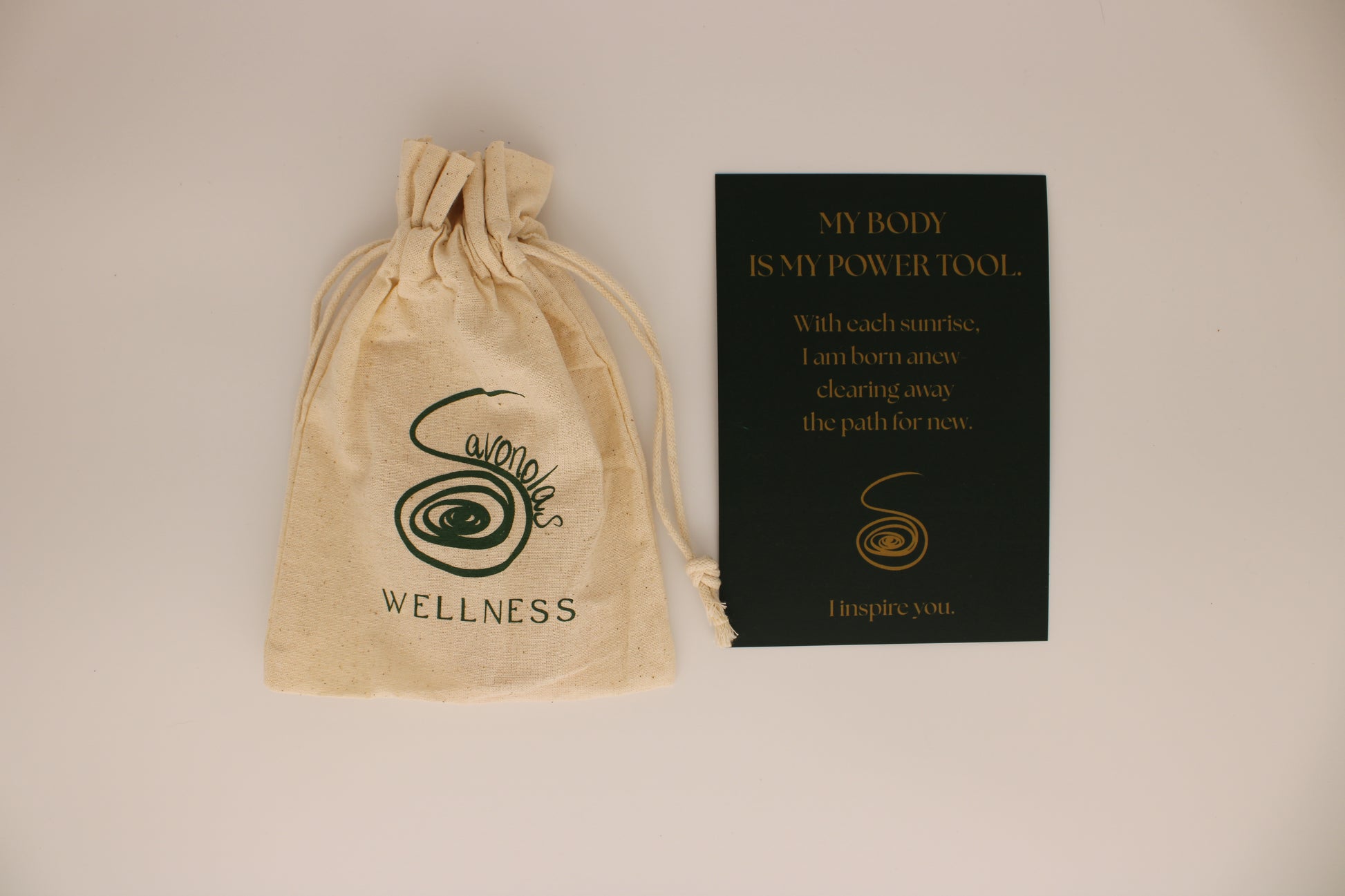 A natural cotton drawstring bag featuring the Savonolas Wellness logo sits beside a deep green affirmation card with gold text. The card reads: “MY BODY IS MY POWER TOOL. With each sunrise, I am born anew—clearing away the path for new. I inspire you.” This packaging reflects the brand’s commitment to intentional wellness and daily ritual enhancement, designed to accompany the copper tongue scraper.