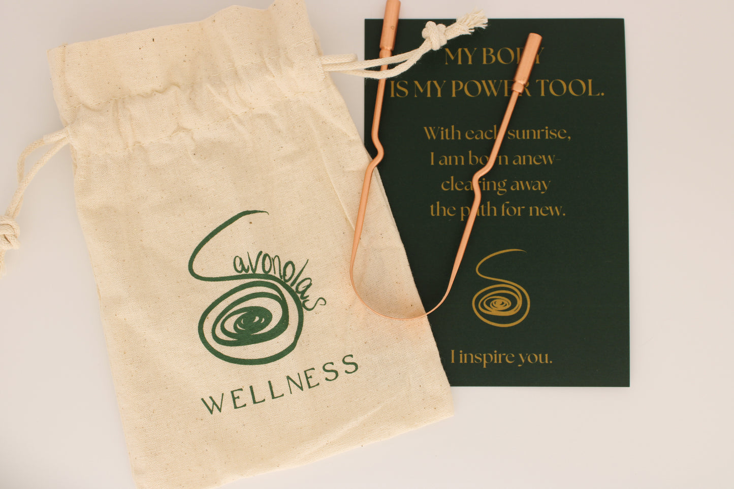This styled product image features the Savonolas copper tongue scraper across a natural cotton wellness bag, highlighting the engraved brand logo. Partially beneath the scraper lies an elegant forest-green affirmation card with golden text that reads: "My body is my power tool. With each sunrise, I am born anew—clearing away the path for new. I inspire you." The image captures the intentional beauty and grounding ritual of using Savonolas wellness tools—merging practicality with spirit