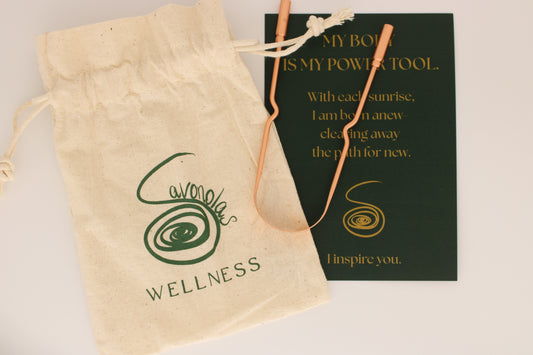 This styled product image features the Savonolas copper tongue scraper across a natural cotton wellness bag, highlighting the engraved brand logo. Partially beneath the scraper lies an elegant forest-green affirmation card with golden text that reads: "My body is my power tool. With each sunrise, I am born anew—clearing away the path for new. I inspire you." The image captures the intentional beauty and grounding ritual of using Savonolas wellness tools—merging practicality with spirit